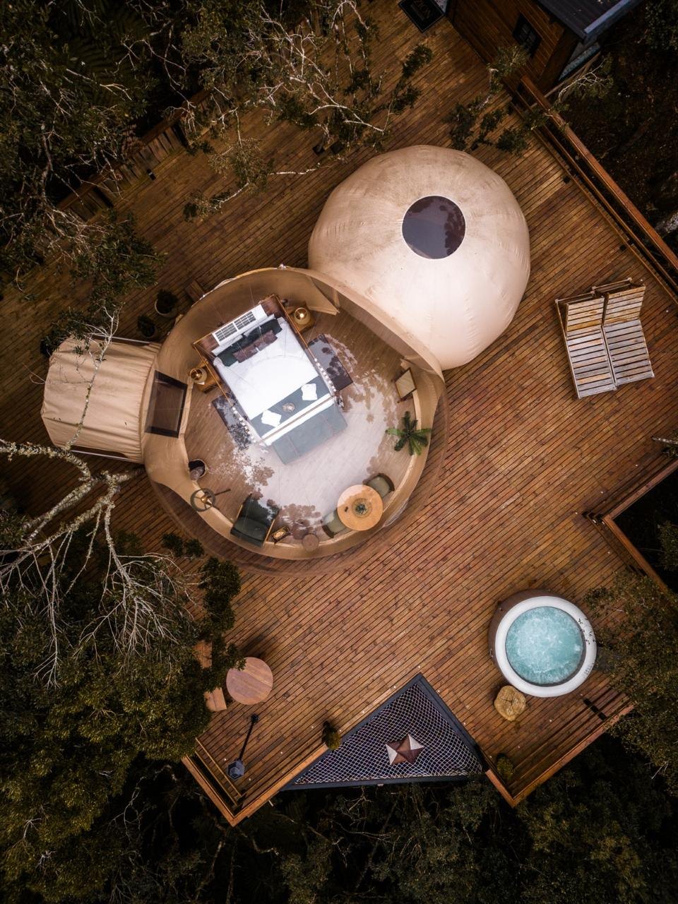 Vista aérea diurna de um bubble Zion sobre deck de madeira, com ofurô e araucárias ao redor